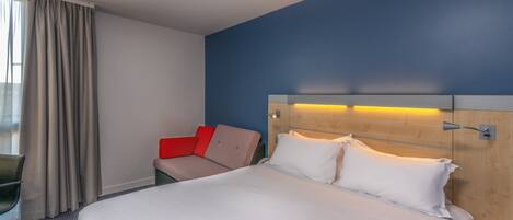 A hotel room with a large bed, a desk, a chair, and a sofa with red cushions.