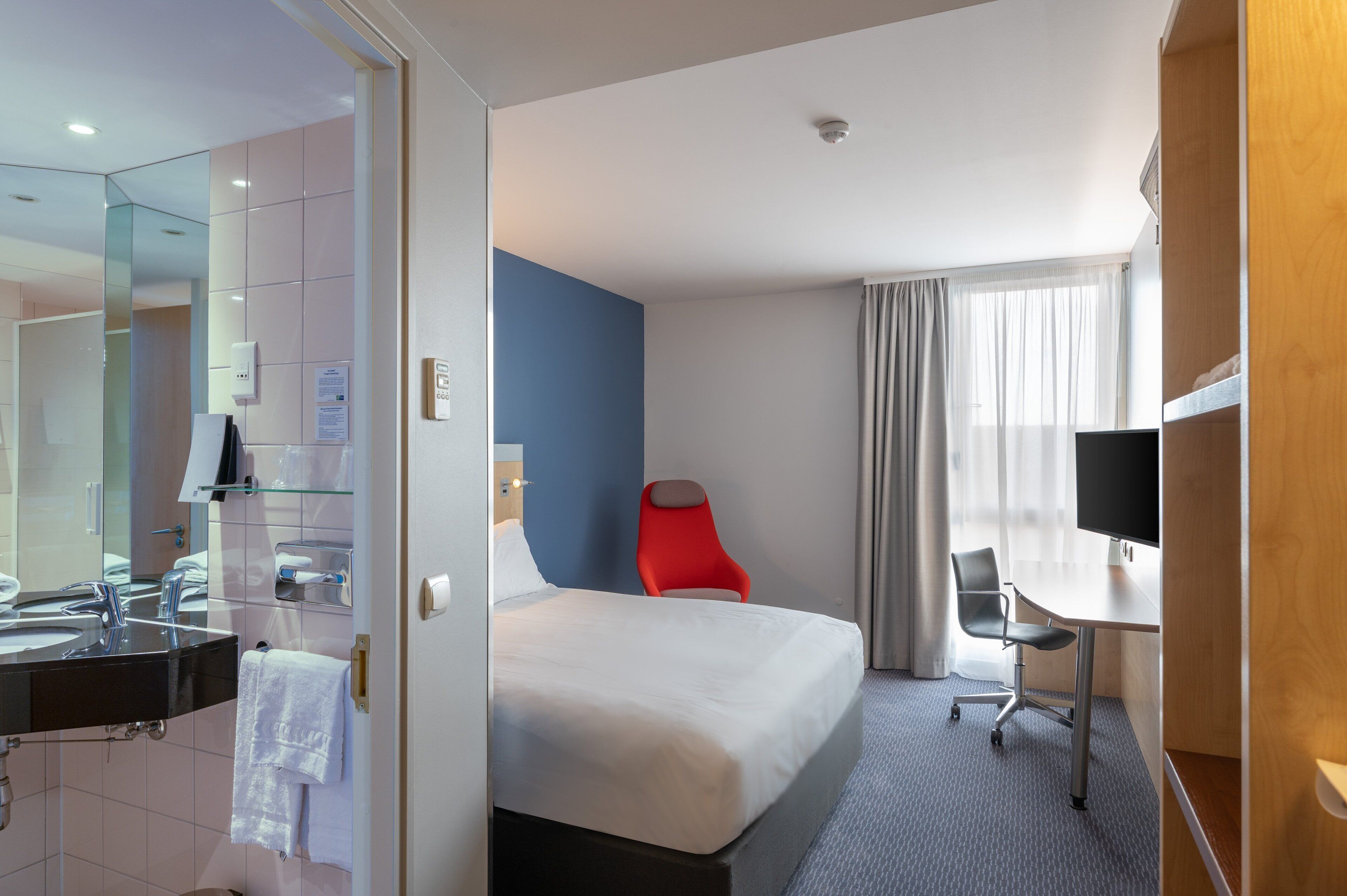 Ett hotellrum med en stor säng, ett skrivbord, en stol, en TV och ett badrum med spegel och handfat.
