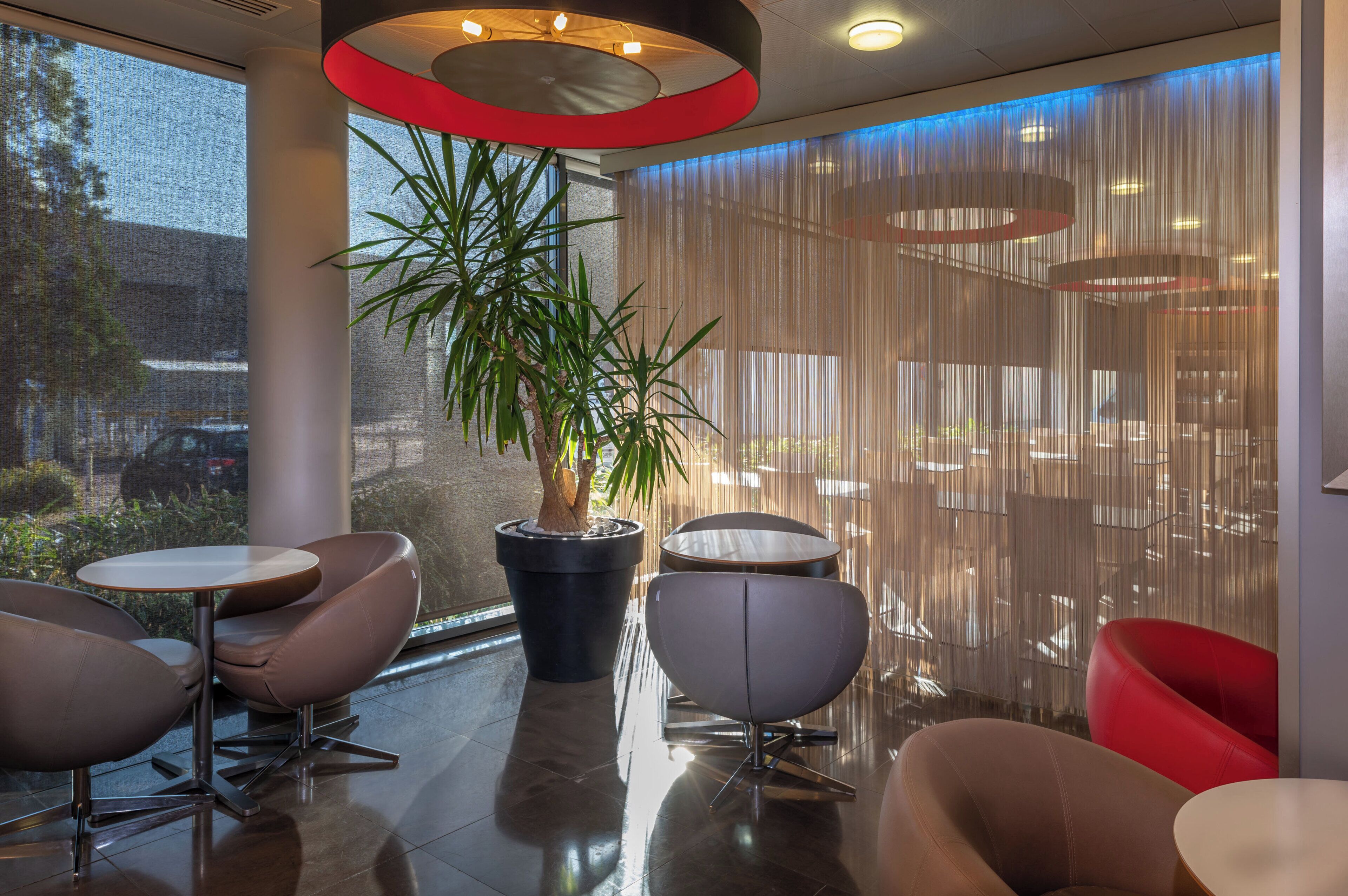 Un espacio interior moderno con una planta en maceta grande, arreglos de asientos y vistas por una ventana.