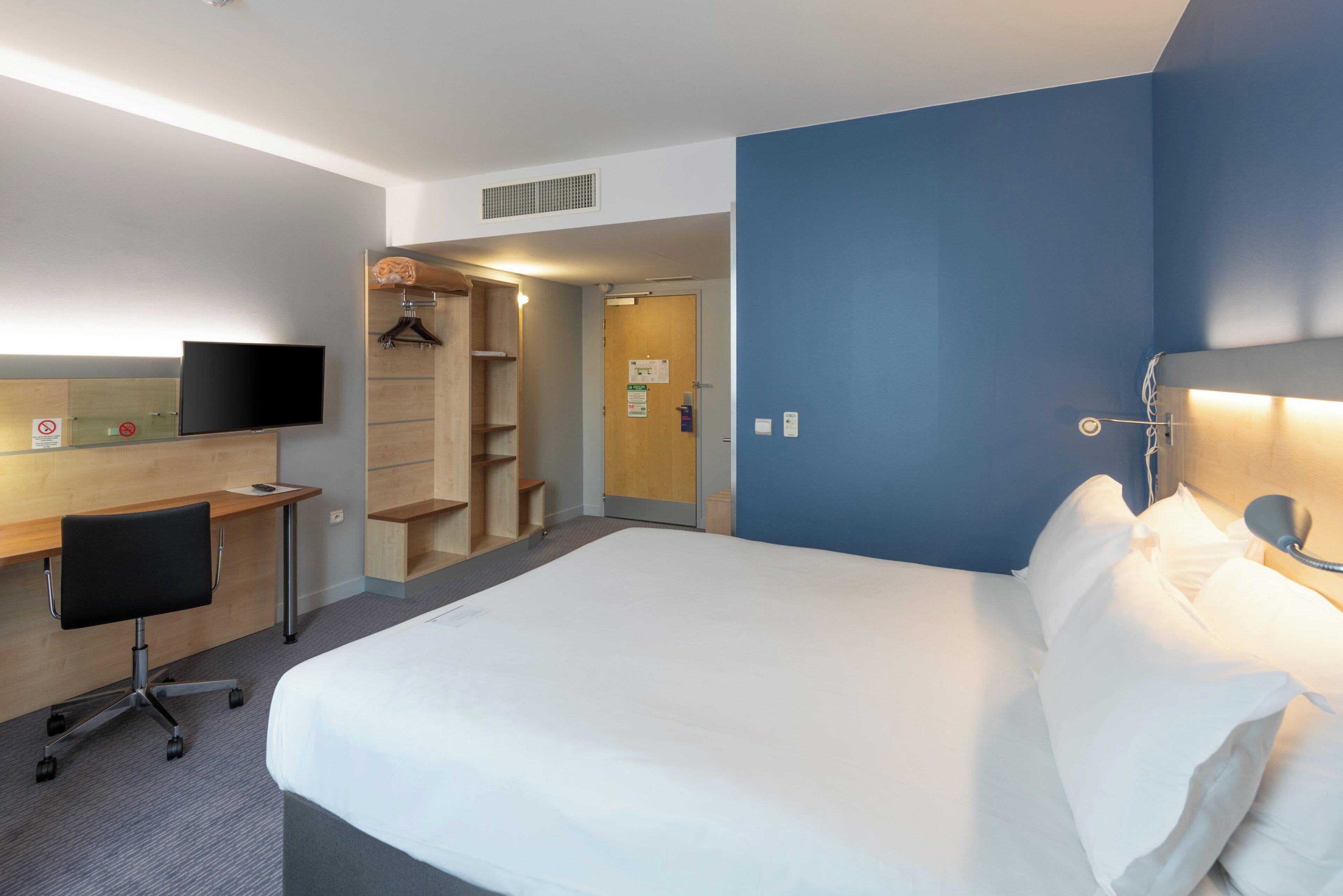 Quarto de hotel com uma cama, uma escrivaninha com cadeira, televisão e guarda-roupa.