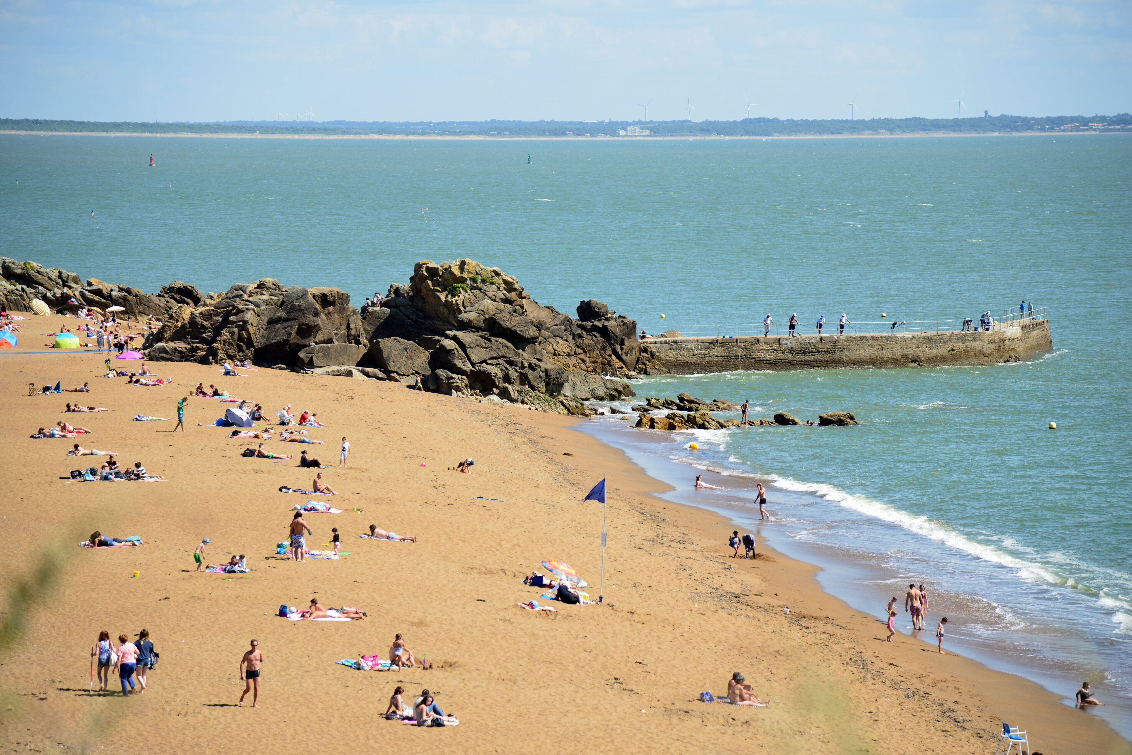 Une plage où des gens se prélassent au soleil et se baignent, un promontoire rocheux et une jetée où se trouvent des personnes.