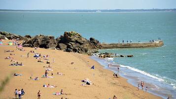 Une plage où des gens se prélassent au soleil et se baignent, un promontoire rocheux et une jetée où se trouvent des personnes.