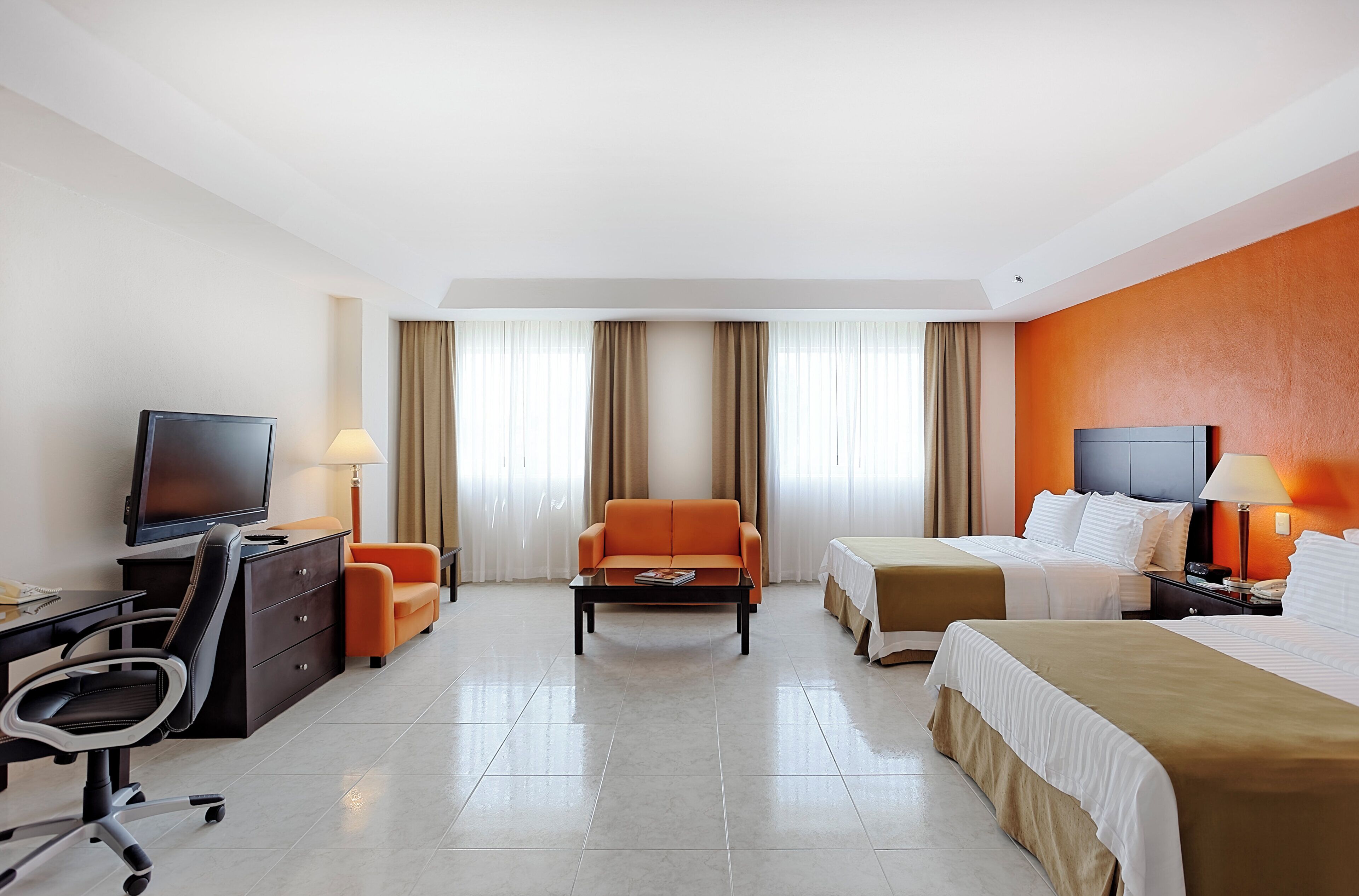 A hotel room with a bed, a sofa, a desk with a computer, a TV, and a window with curtains.