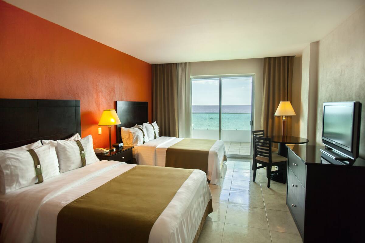 A hotel room with two beds, a TV, a desk, and a view of the ocean.