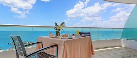 A balcony with a table set for dining, overlooking a clear blue sea and boats.