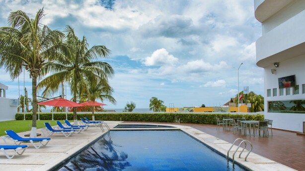 Una piscina con azulejos azules, rodeada de sillas de descanso y palmeras.