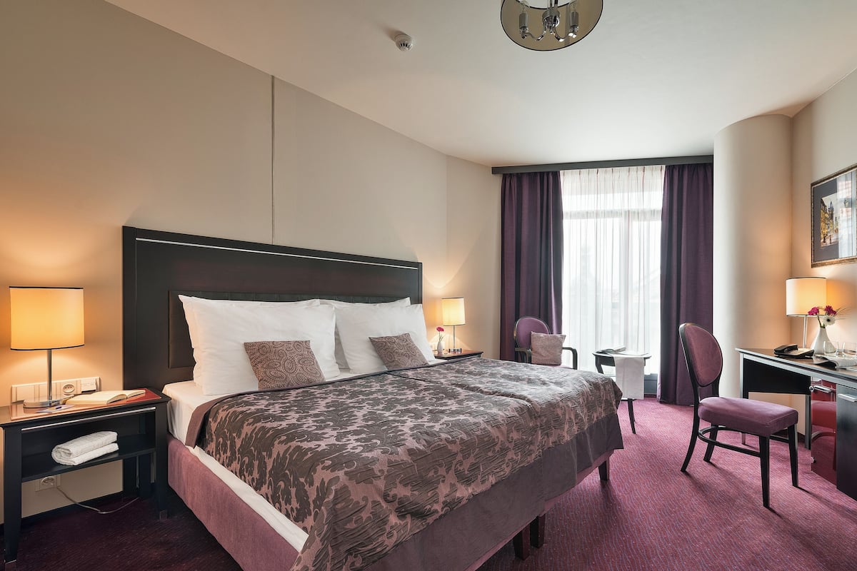 A hotel room with a large bed, two bedside lamps, a desk with a chair, and a window with curtains.