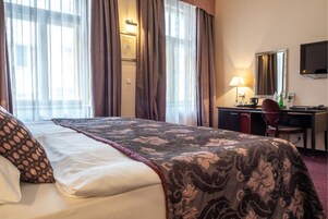 Quarto de hotel com cama, escrivaninha, cadeira, luminária e televisão na parede.
