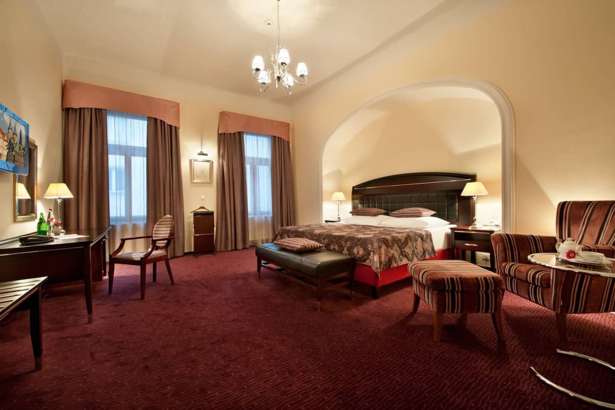 A hotel room with a large bed, a desk, a chair, a bench, and a small table with a teapot.