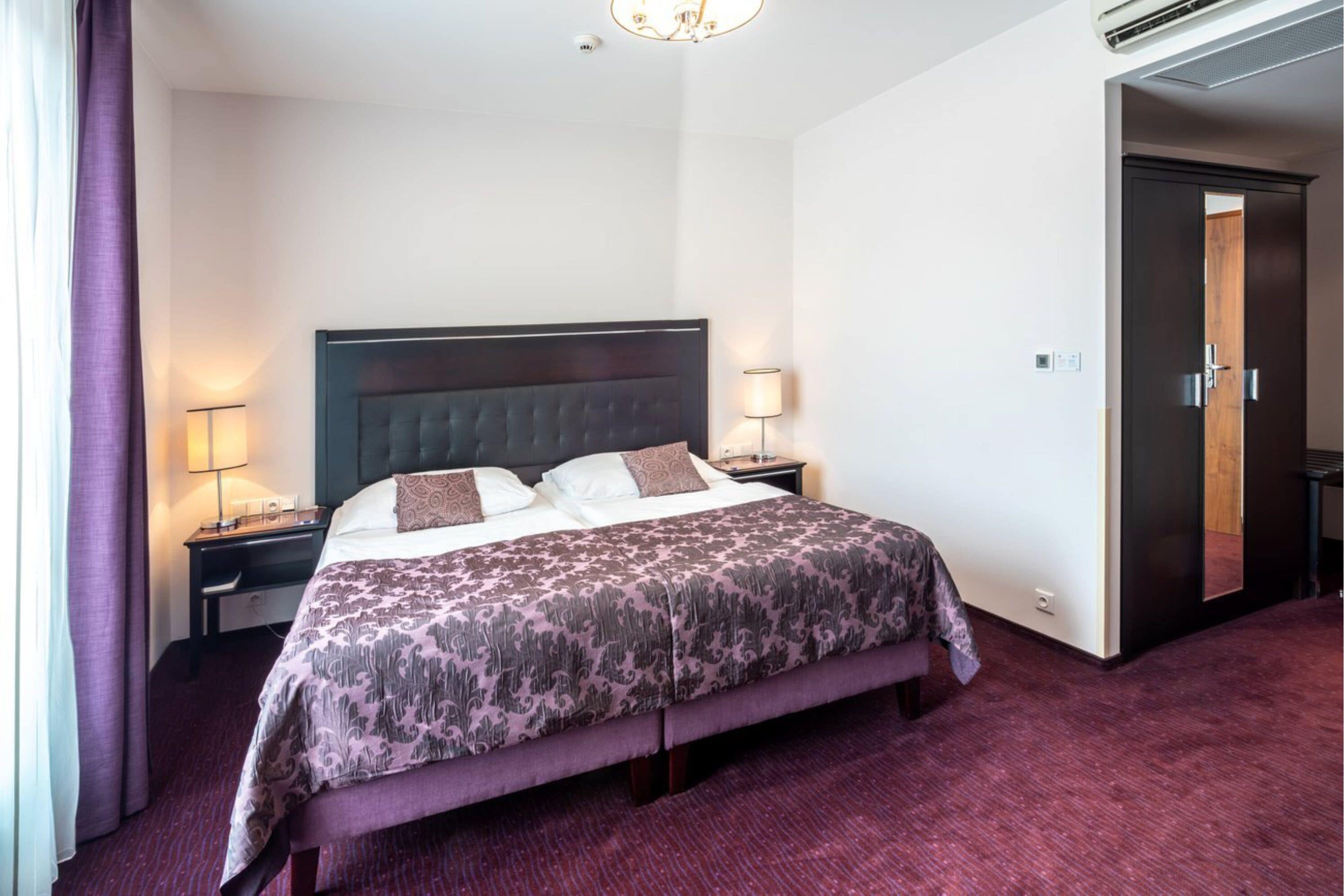 Habitación de hotel con una cama grande, una mesita de noche, una lámpara y una puerta que da a otra habitación.