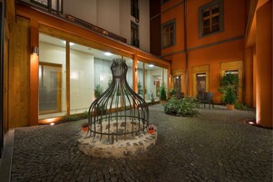 Um pátio com uma escultura metálica única, pavimento de pedras e um edifício com janelas e portas amplas.