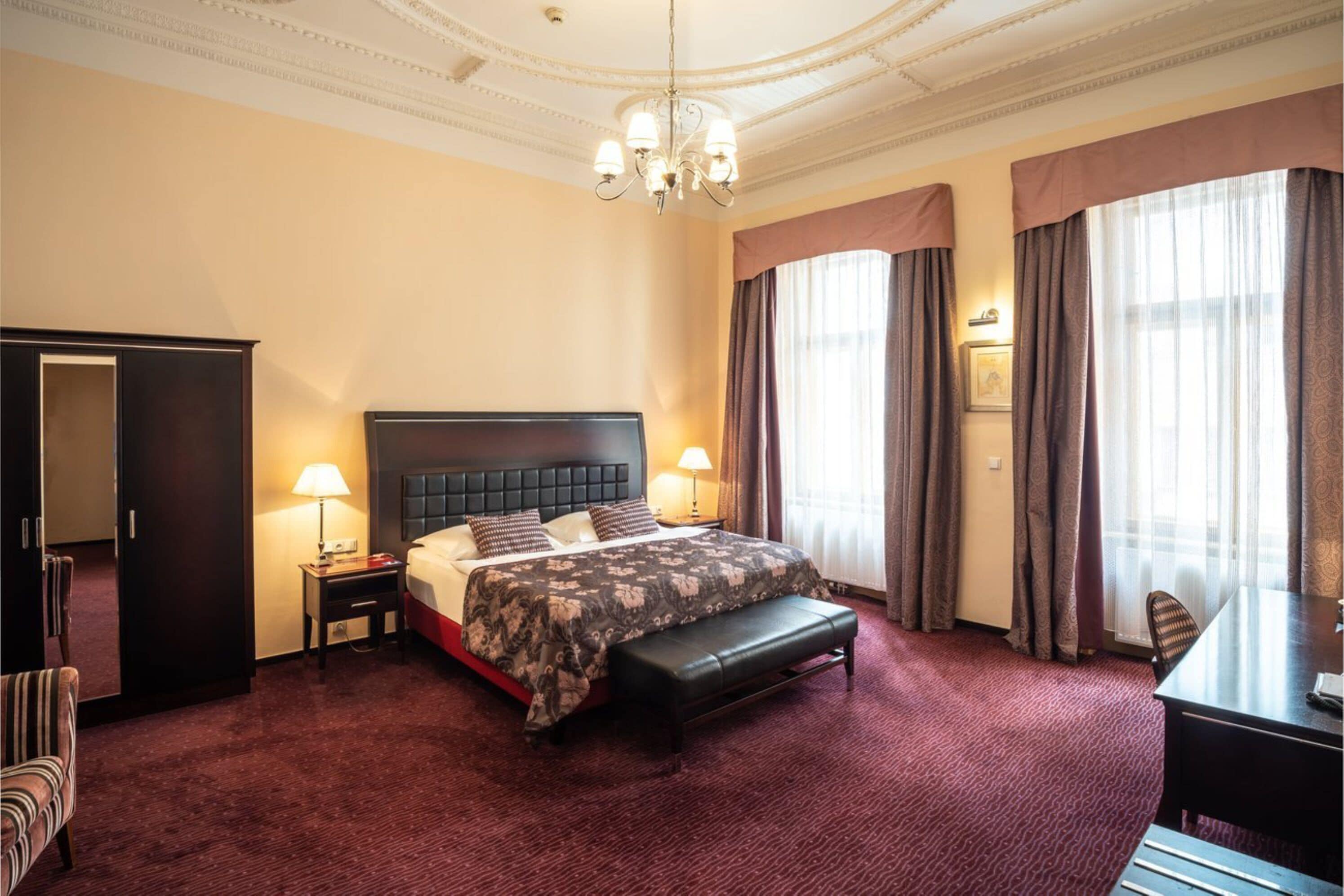 A hotel room with a large bed, a desk, a chair, a wardrobe, and a chandelier.