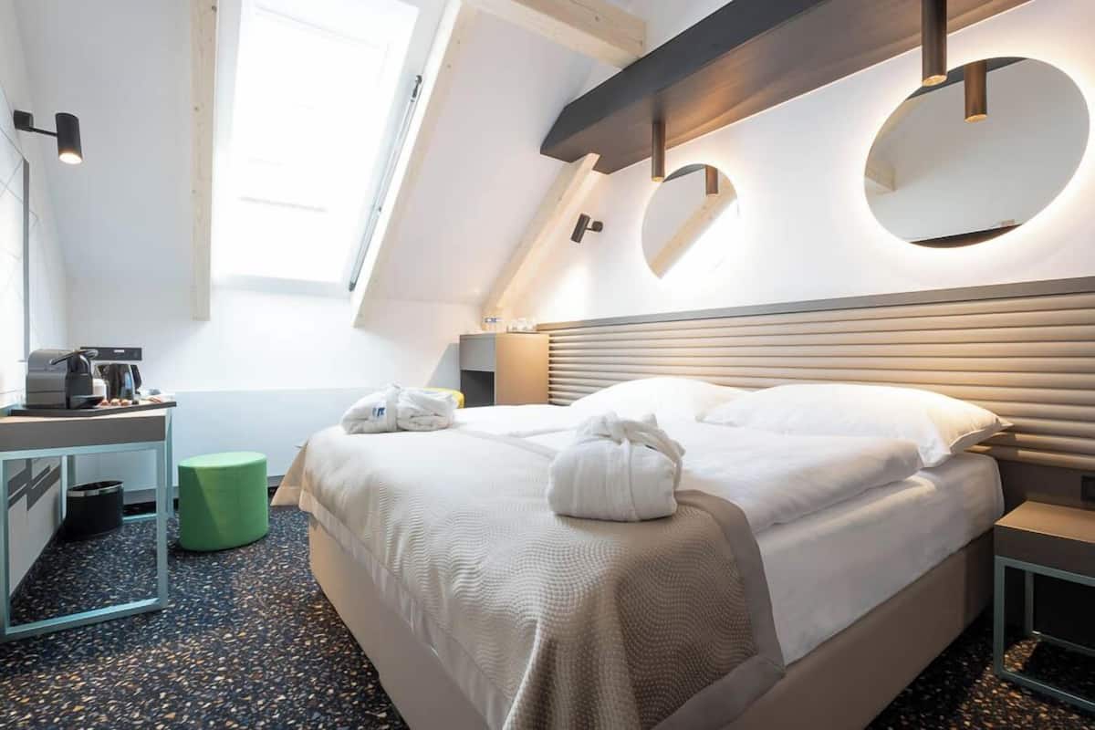 A modern hotel room with a large bed, a desk with a coffee machine, a green stool, and a skylight.