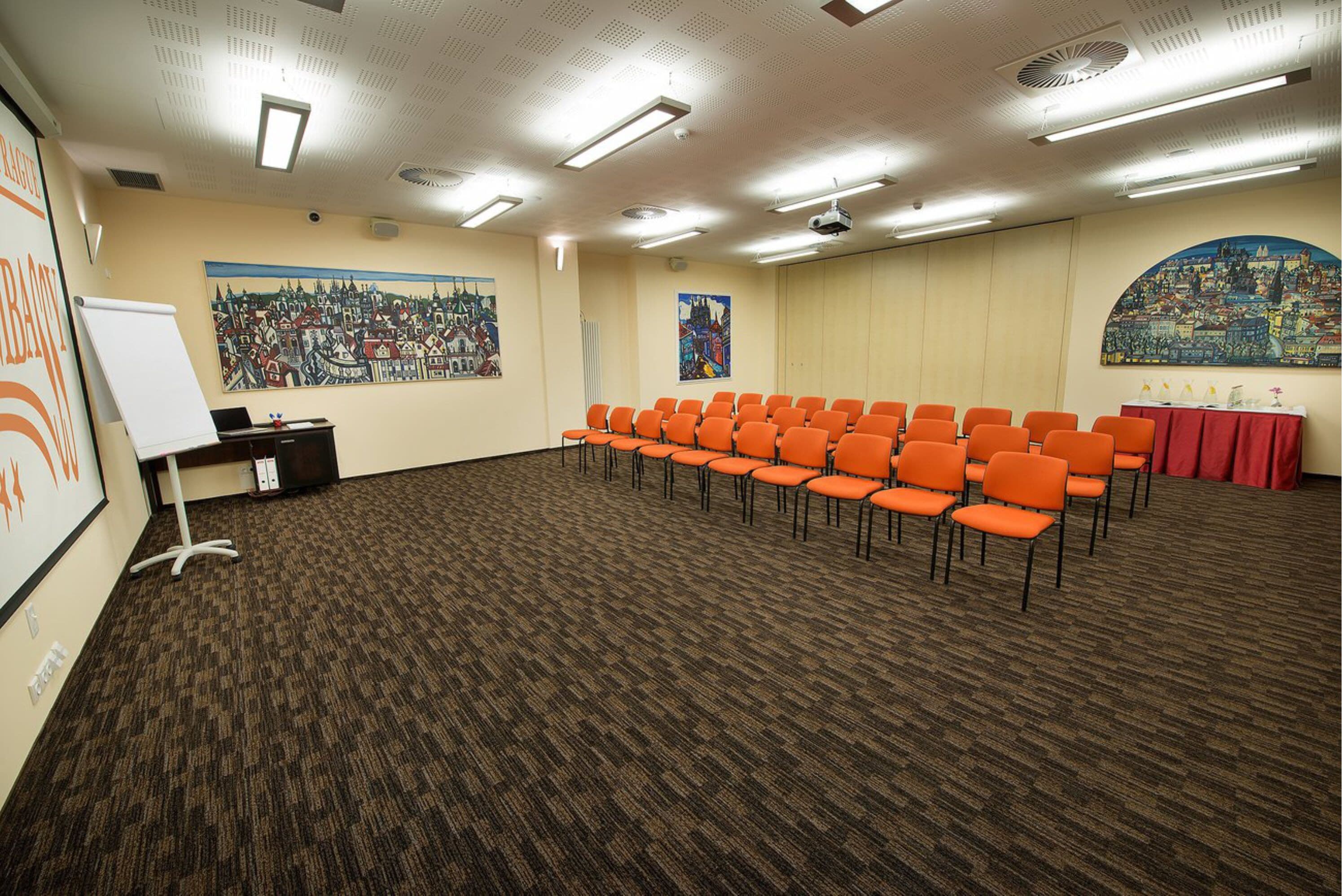 A conference room with rows of orange chairs, a whiteboard, and two large paintings on the walls.