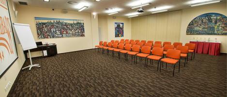 A conference room with rows of orange chairs, a whiteboard, and two large paintings on the walls.