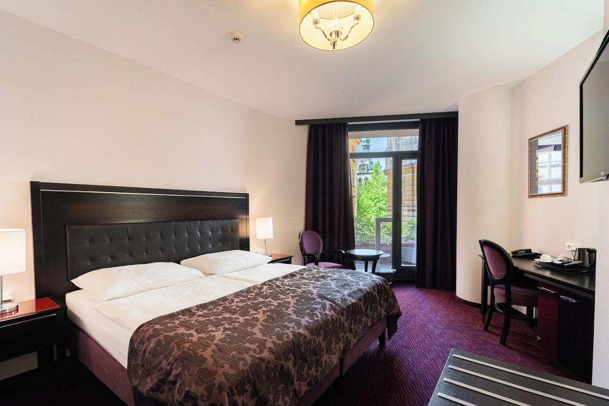 Hotellihuone, jossa on sänky, työpöytä, tuoli, lamppu ja ikkunasta avautuva näkymä ulos.
