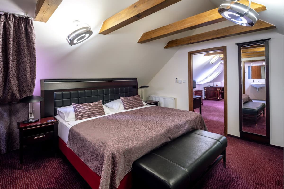 A bedroom with a large bed, a bedside table, a lamp, a bench, and a view of the bathroom through an open door.