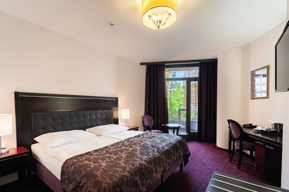 A hotel room with a bed, a desk, a chair, a lamp, and a view of the outside.