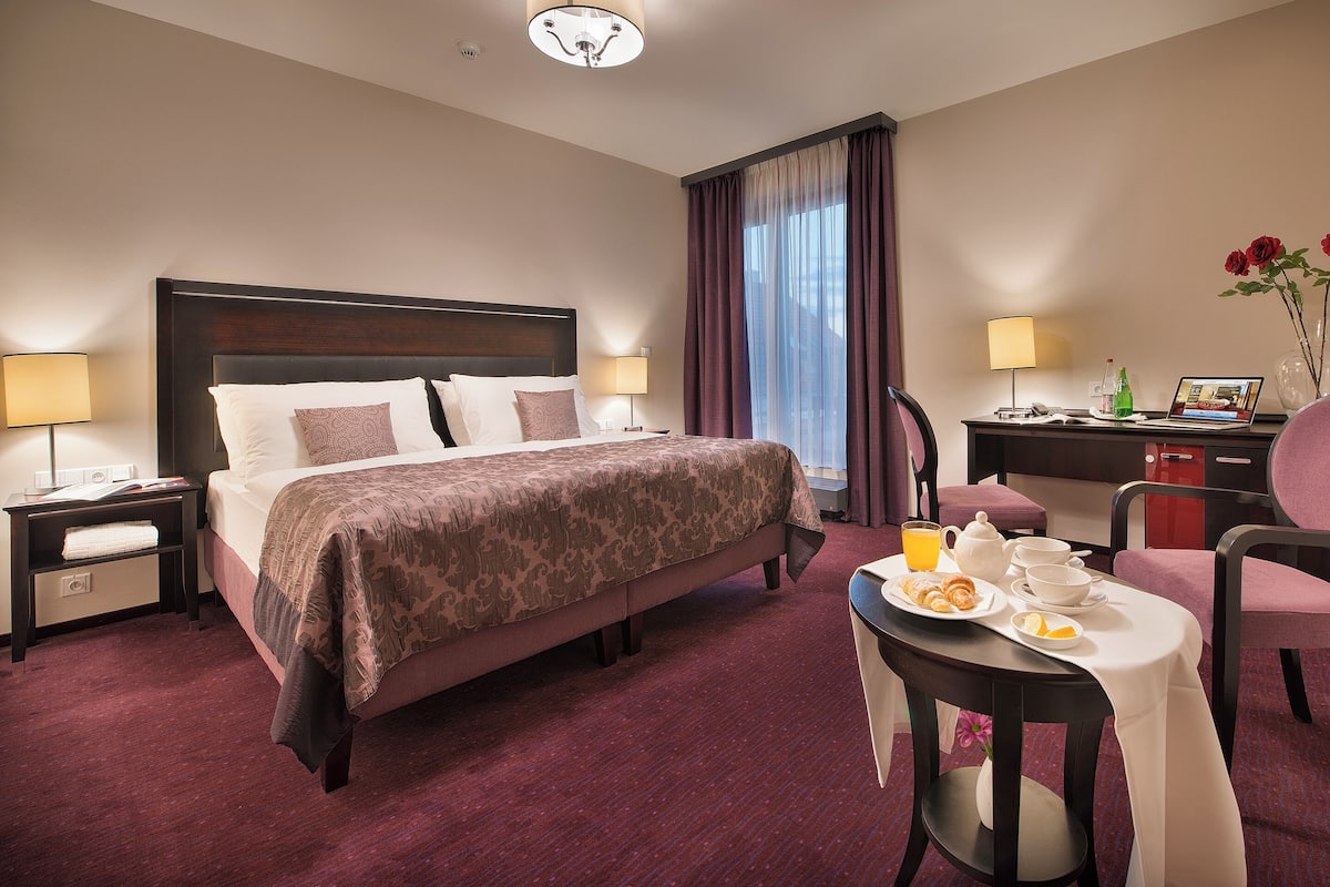 A hotel room with a large bed, a desk with a laptop, a chair, and a table with breakfast items.