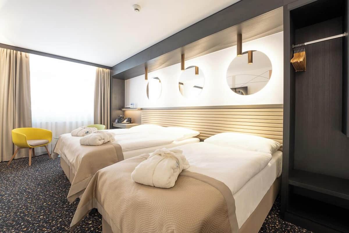 A hotel room with two beds, a yellow chair, a mirror, and a window with curtains.