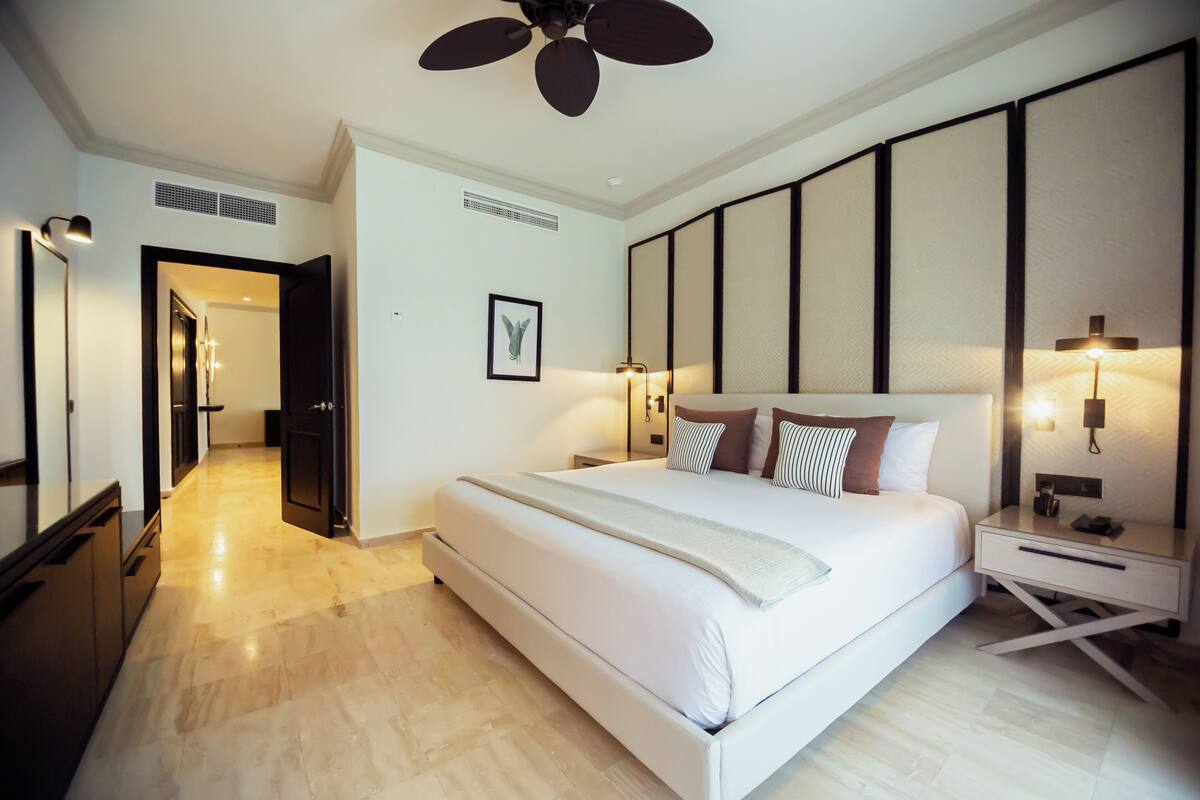 A modern hotel room with a large bed, bedside tables, a ceiling fan, and a view of the hallway.