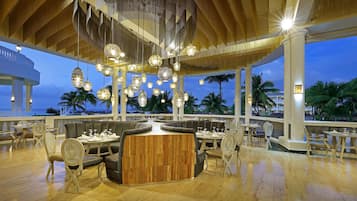 Un vaste espace extérieur aménagé pour les repas, avec de nombreux coins salon, des guirlandes lumineuses décoratives et une vue sur l’océan.