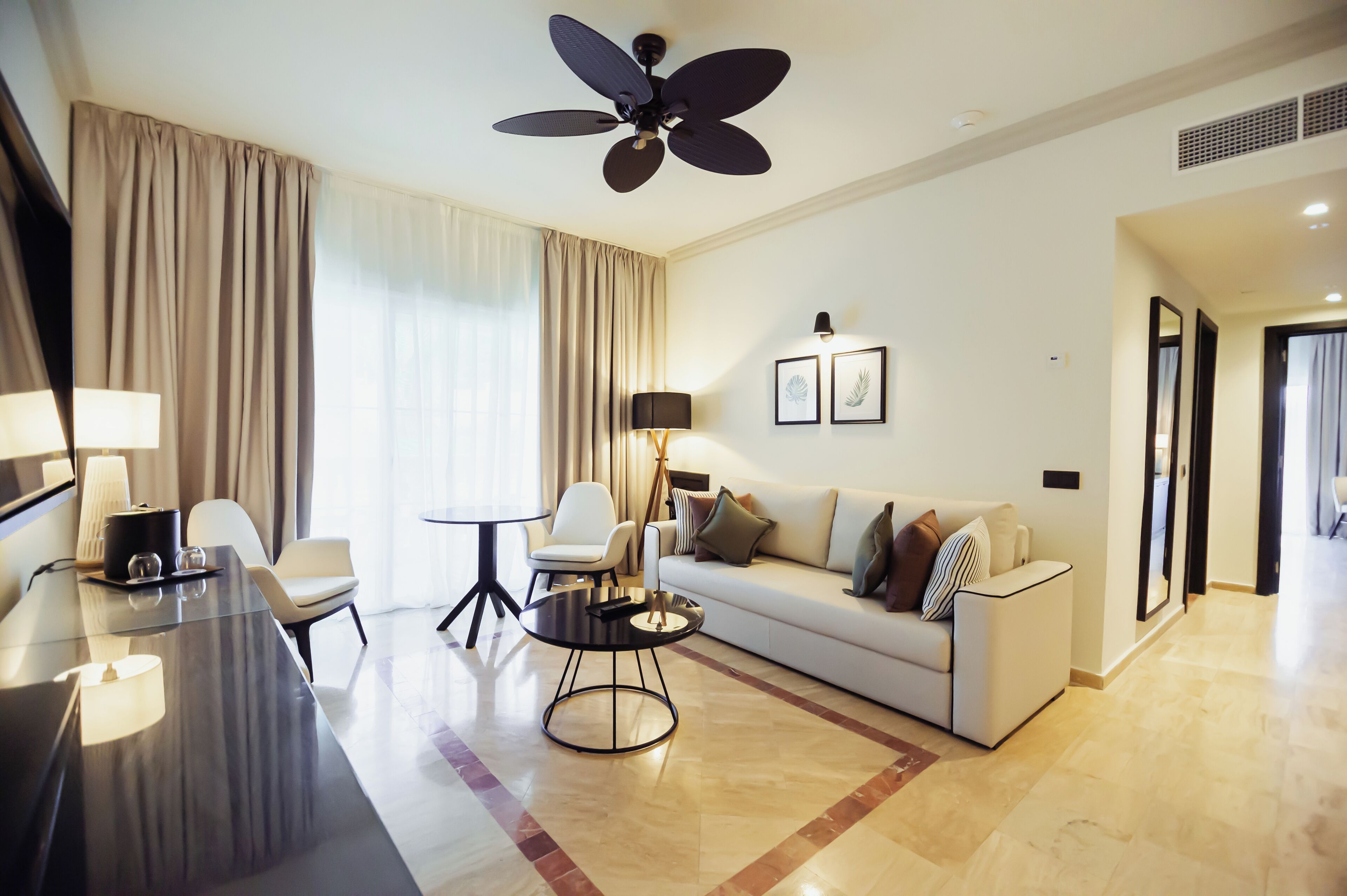 A modern hotel room with a large ceiling fan, a sofa, a dining table, and a lamp.