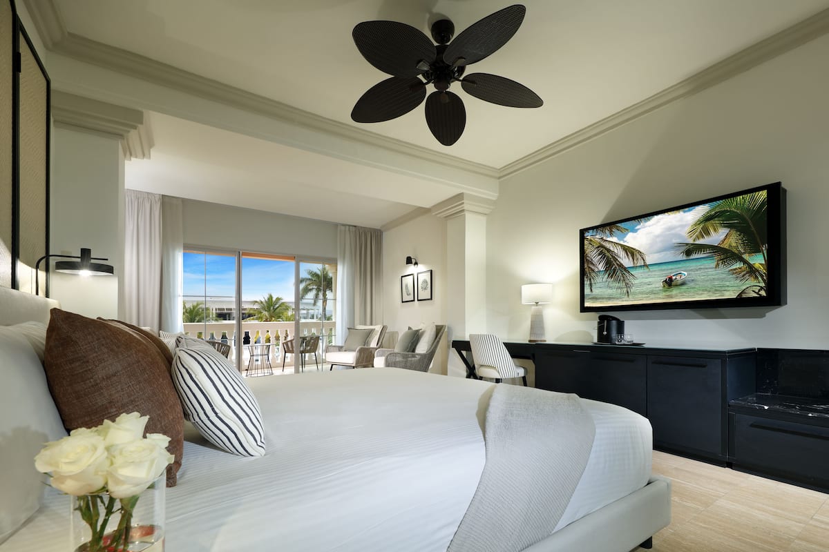 A hotel room with a large bed, a flat-screen TV, a ceiling fan, and a view of the ocean through the window.