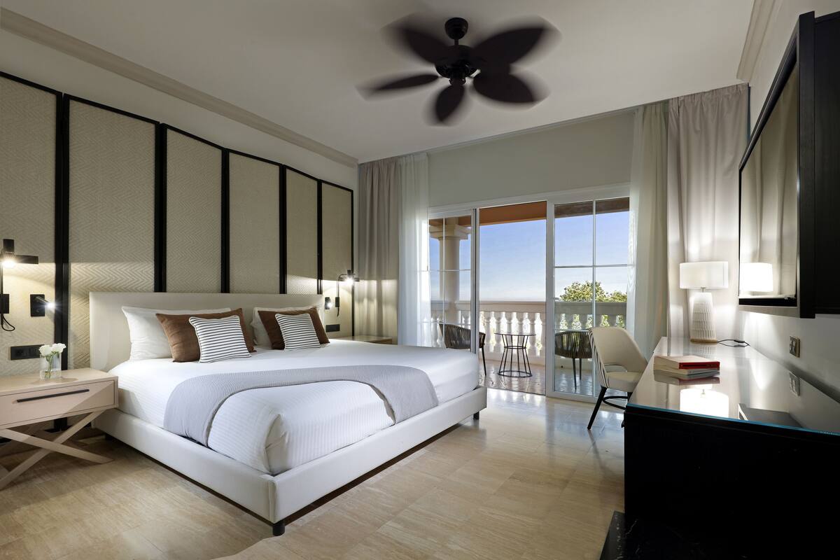 A modern hotel room with a large bed, a desk, a chair, a balcony with a view, and a ceiling fan.