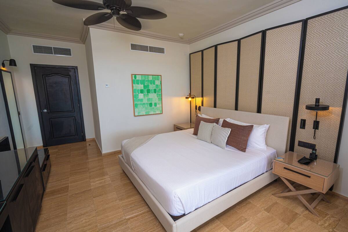 A modern hotel room with a large bed, a bedside table with a lamp, a ceiling fan, and a decorative screen.
