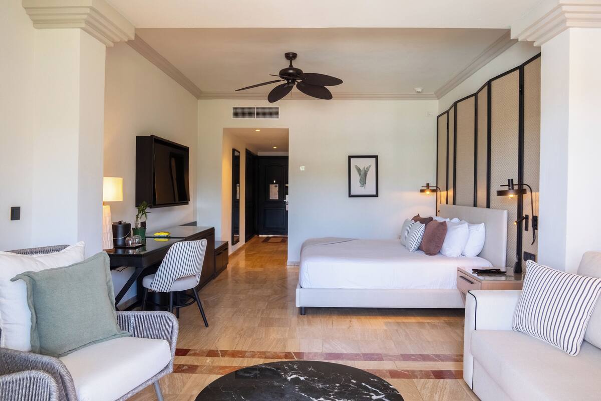 A hotel room with a bed, two white sofas, a dining table, a TV, and a ceiling fan.