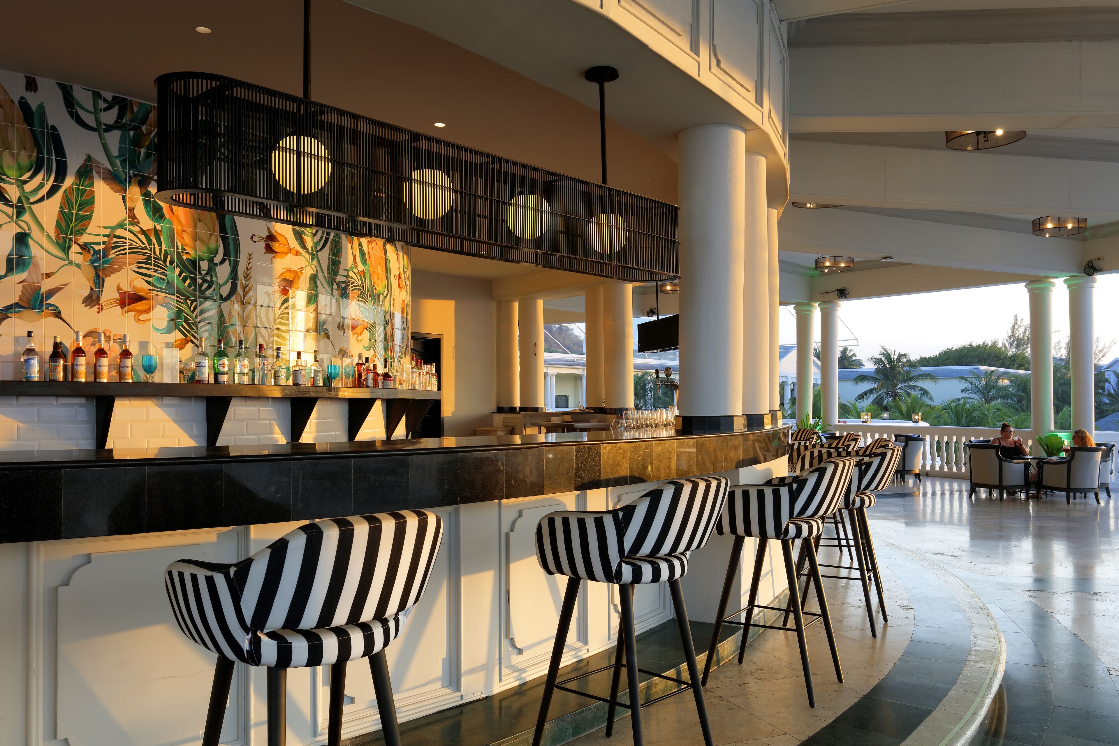 Un área de bar con taburetes a rayas, un mural con hojas tropicales y vistas a palmeras.