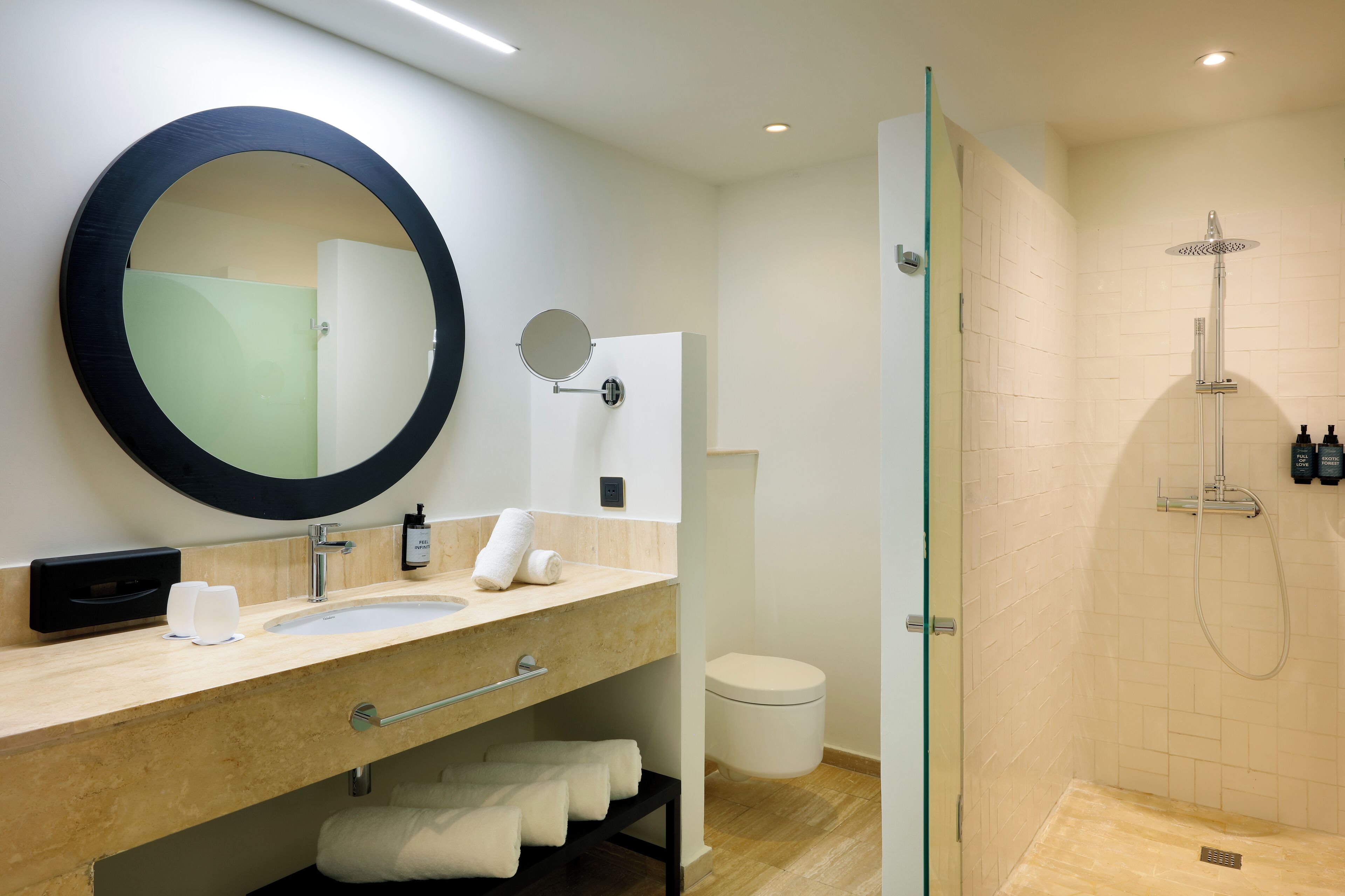 A modern bathroom with a large round mirror, a sink, a glass-enclosed shower, and neatly arranged towels.