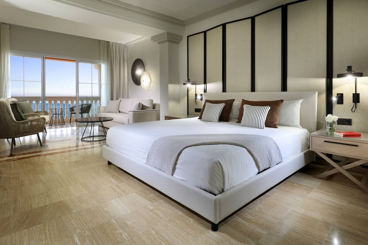 A spacious bedroom with a large bed, a seating area with a chair and table, and a balcony with a view.