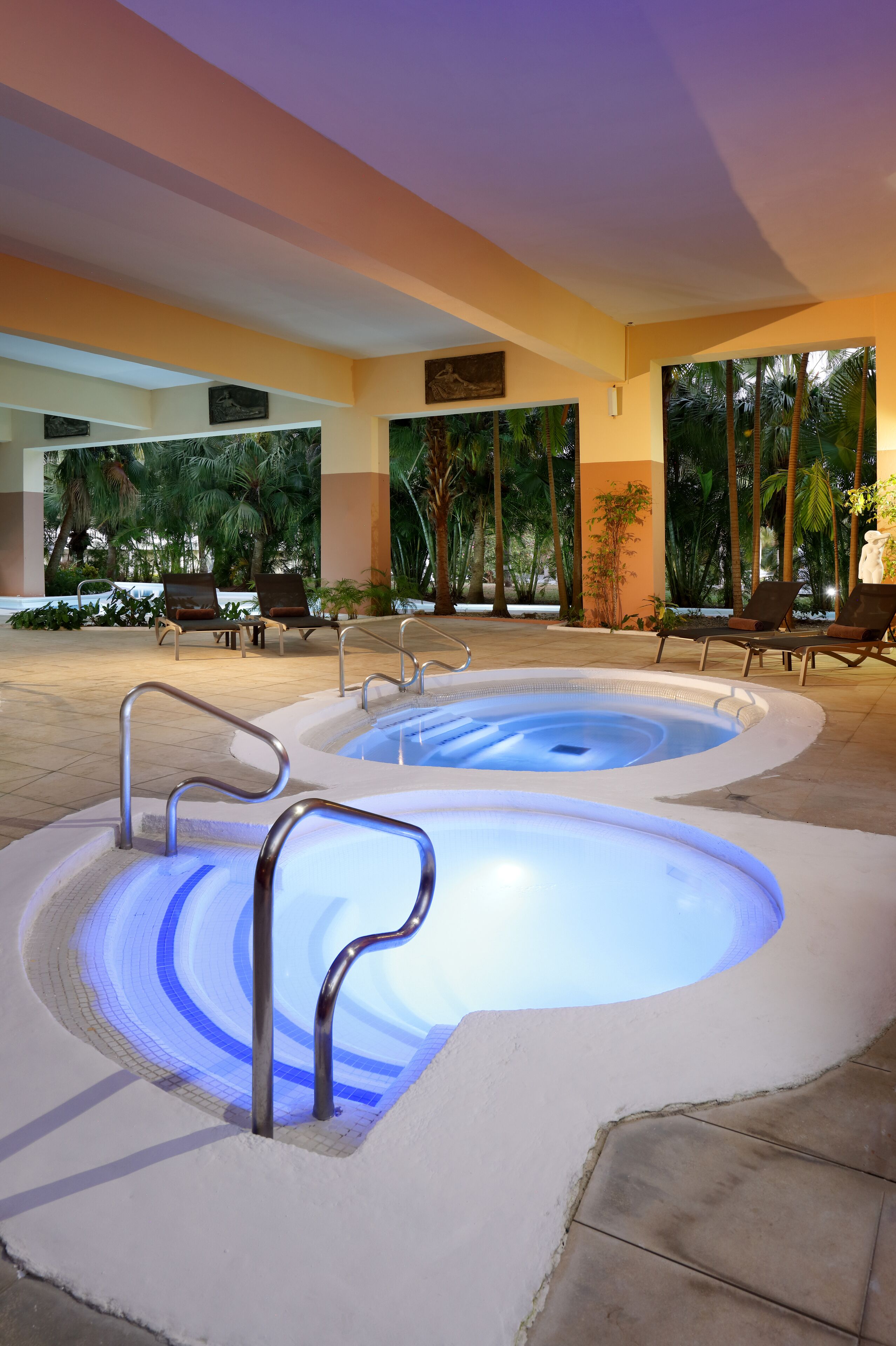 A heated indoor pool area with lounge chairs and palm trees visible through the windows.