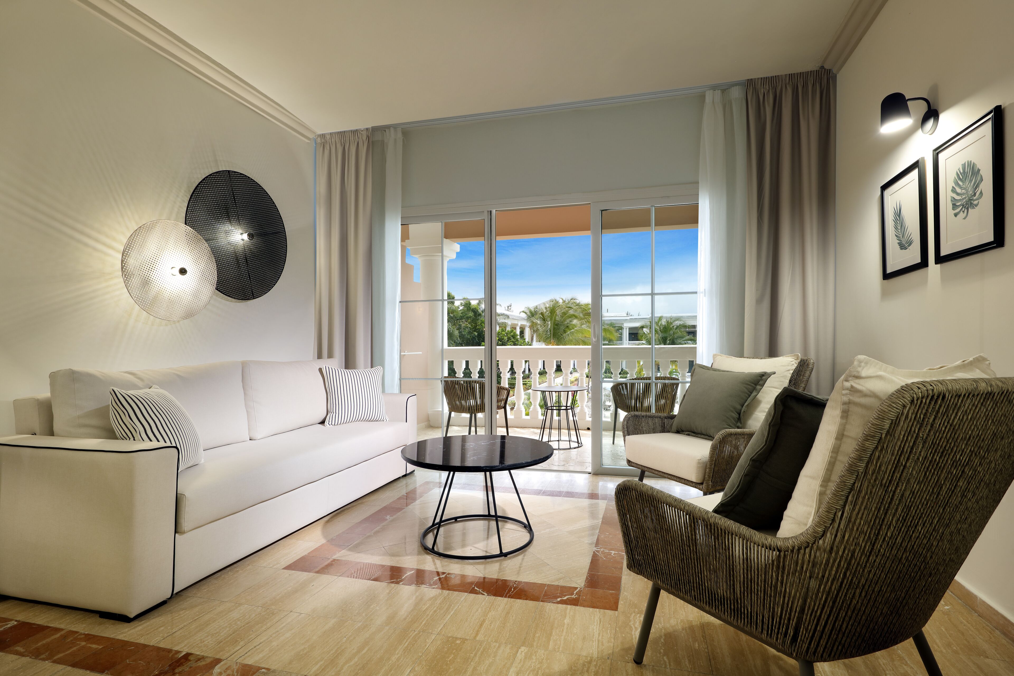 A modern living room with a white sofa, wicker armchair, and a round coffee table. Large windows with curtains provide a view of the outdoors.