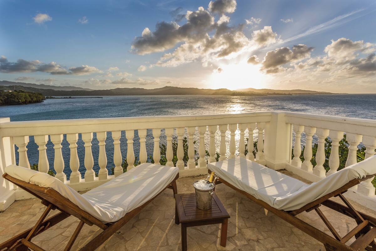 A balcony with two wooden lounge chairs and a small table, overlooking a body of water with a distant shoreline and mountains.