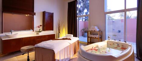 A modern spa room with a large bathtub, a massage table, and a large window with a view of palm trees.