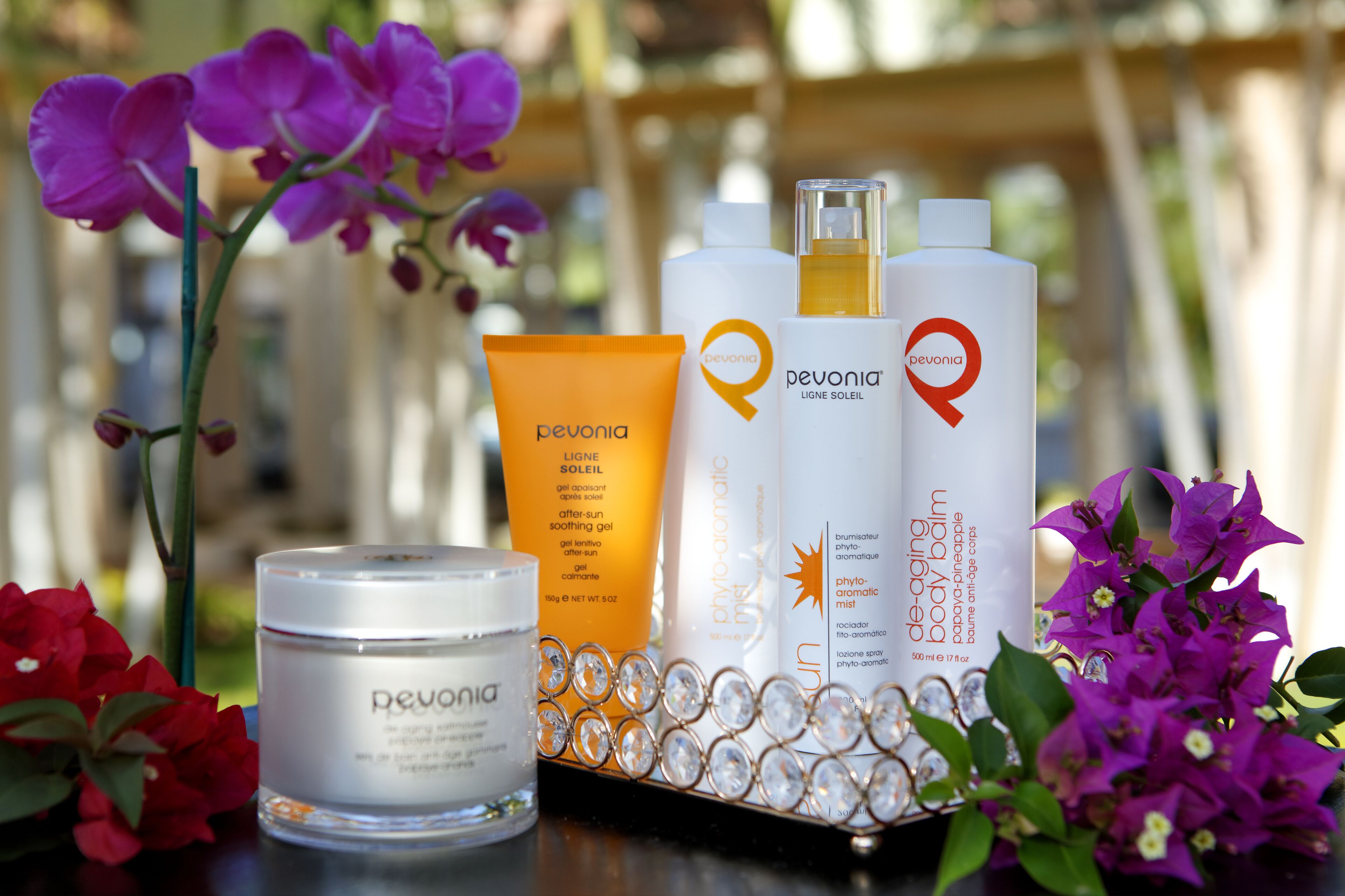 A display of Pevonia skincare products including a jar, a tube, and two bottles, surrounded by flowers.