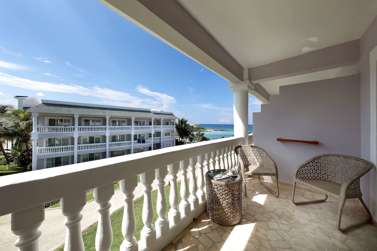 A balcony with a white balustrade, wicker chairs, a small round table with a cup and saucer, and a view of a large building and the ocean.