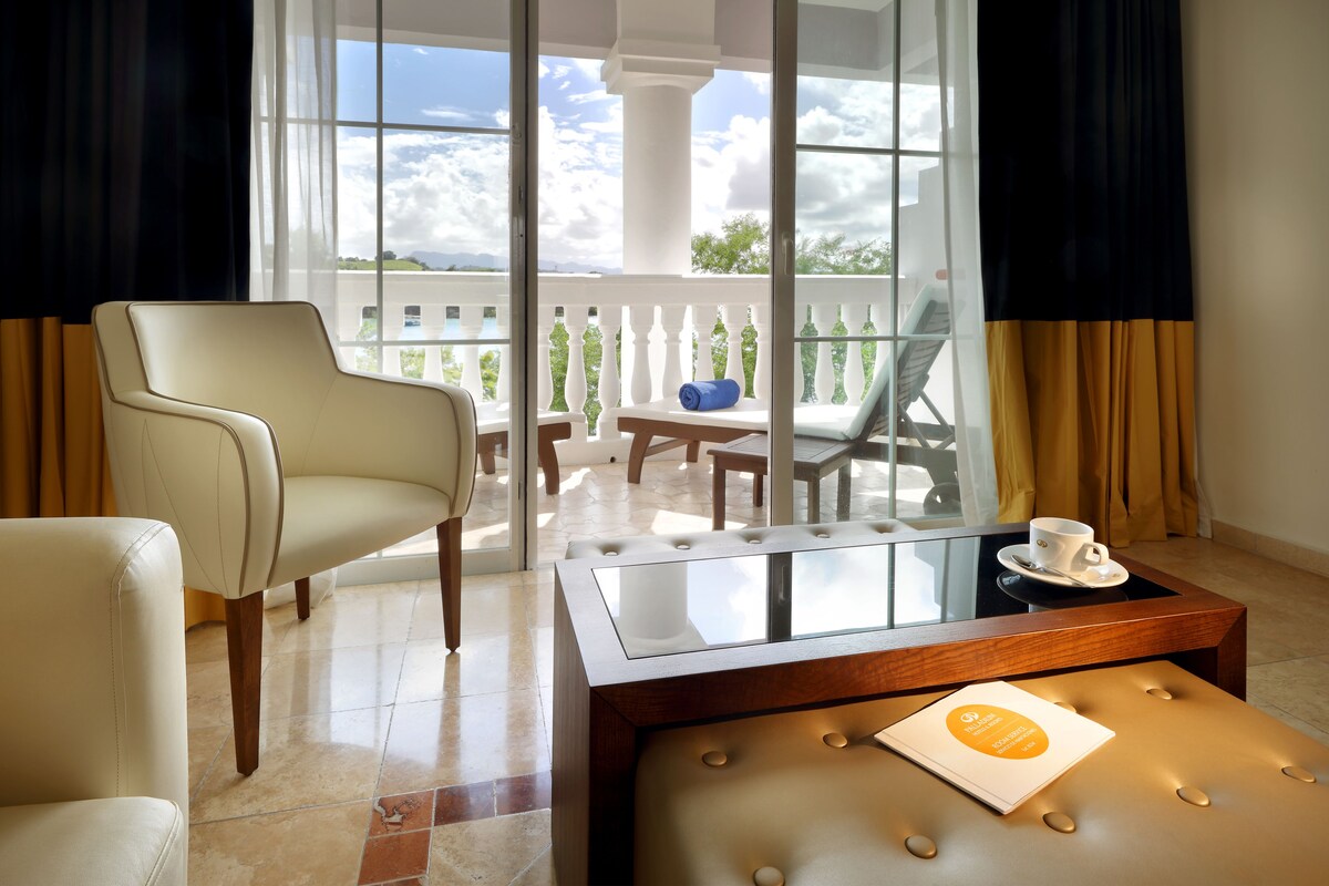 A modern room with a large window, a cushioned bench, a chair, and a small table with a cup and a booklet.