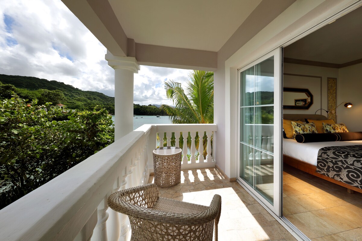 A balcony with wicker furniture, a glass door leading to a bedroom with a patterned bedspread, and a view of a lush green landscape and water.