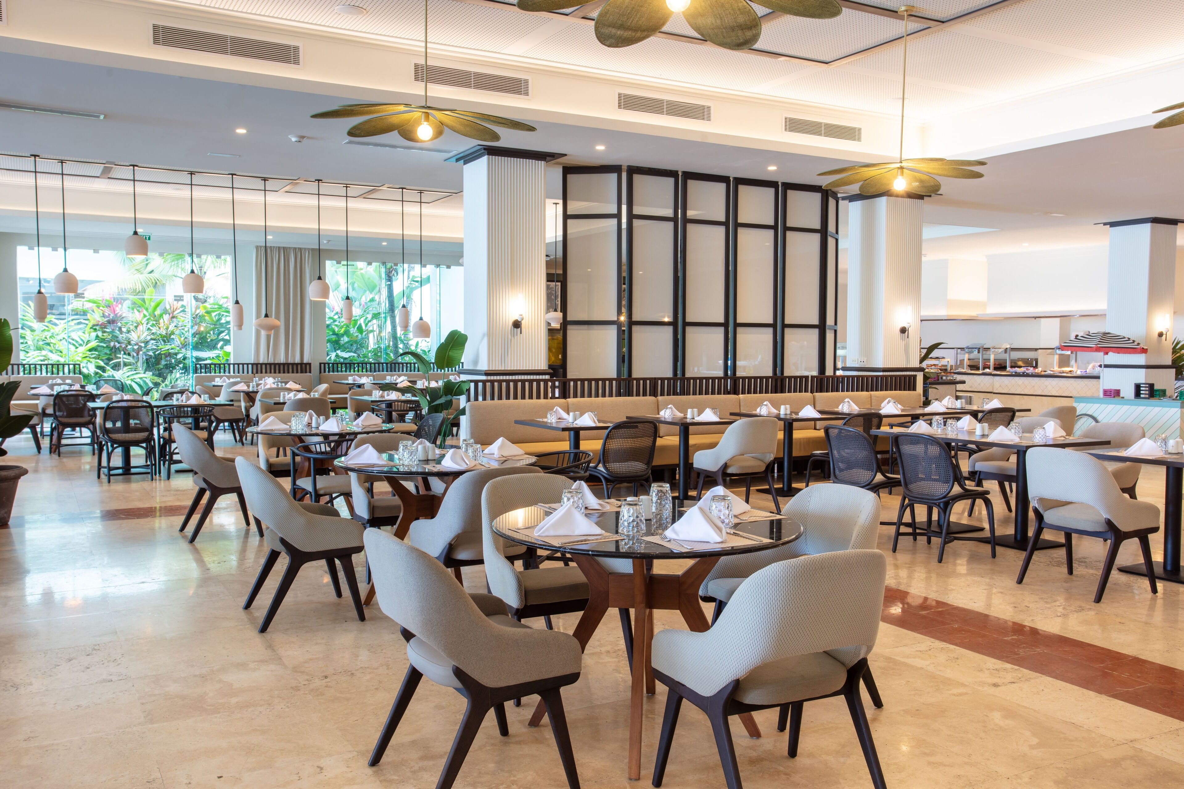 A spacious dining area with multiple tables and chairs, large windows offering views of greenery, and a modern ceiling design.