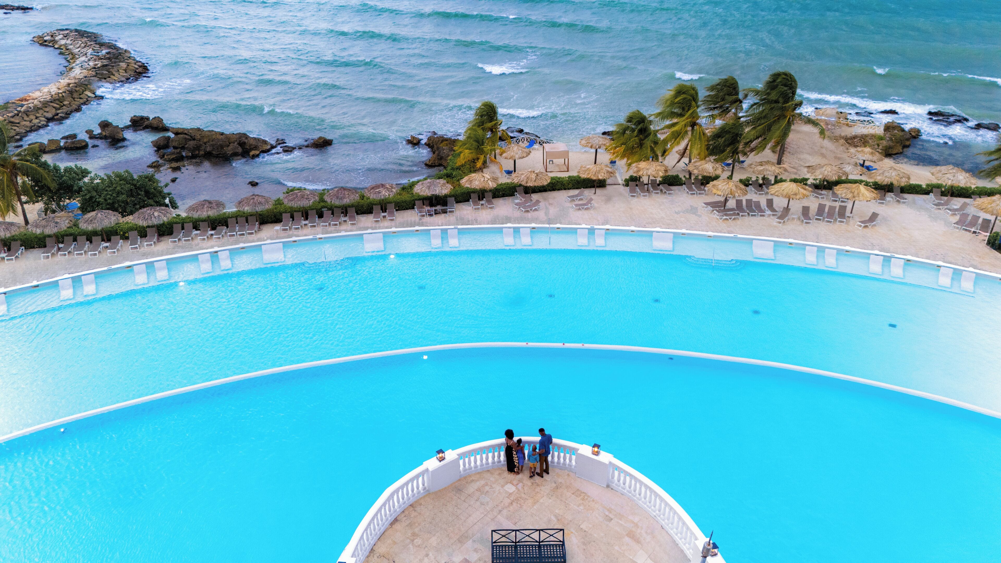 A large, curved swimming pool with a white deck and a few people standing at the edge.