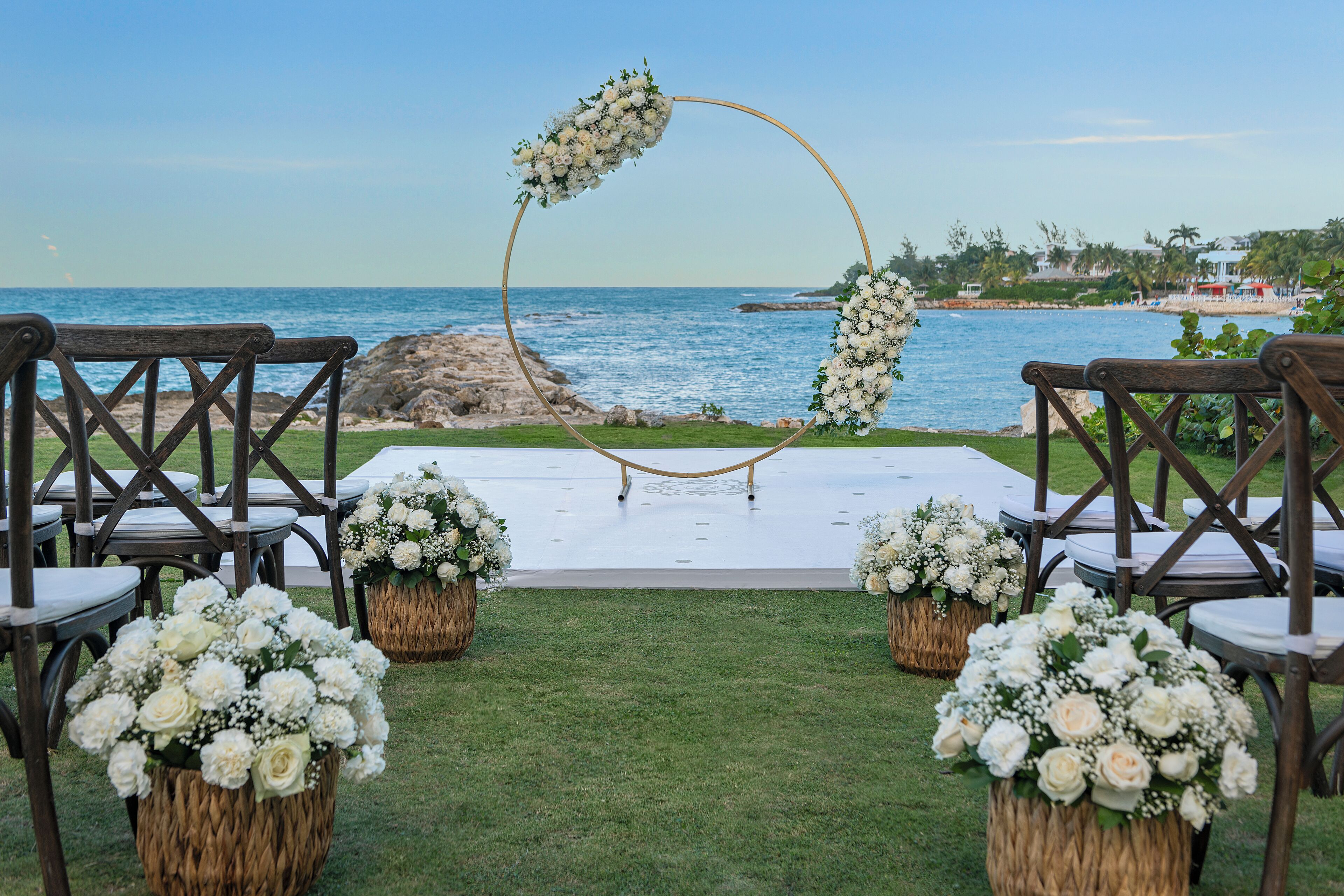 A beachside ceremony setup with a white aisle, wooden chairs, and floral arrangements.