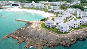 An aerial view of a coastal resort with multiple white buildings, a sandy beach, and clear turquoise waters.