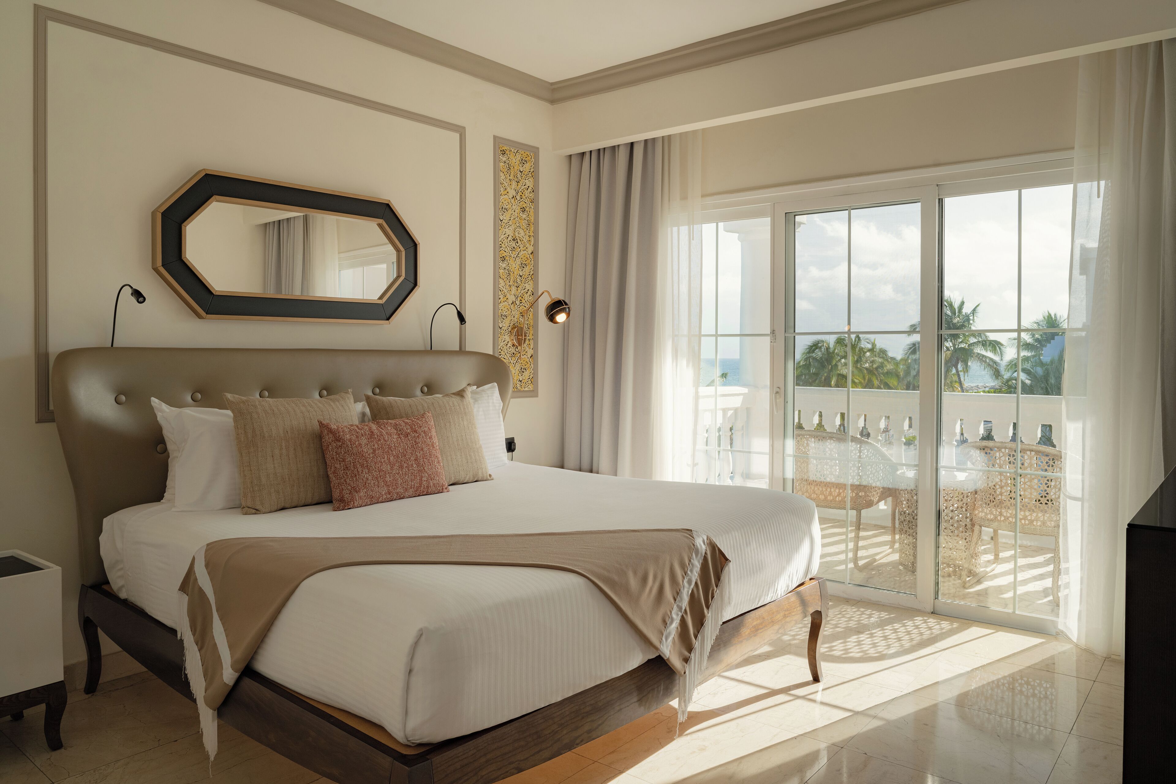 Une chambre spacieuse avec un grand lit, une vue sur l’extérieur grâce à une porte-fenêtre coulissante, et un miroir décoratif au-dessus du lit.