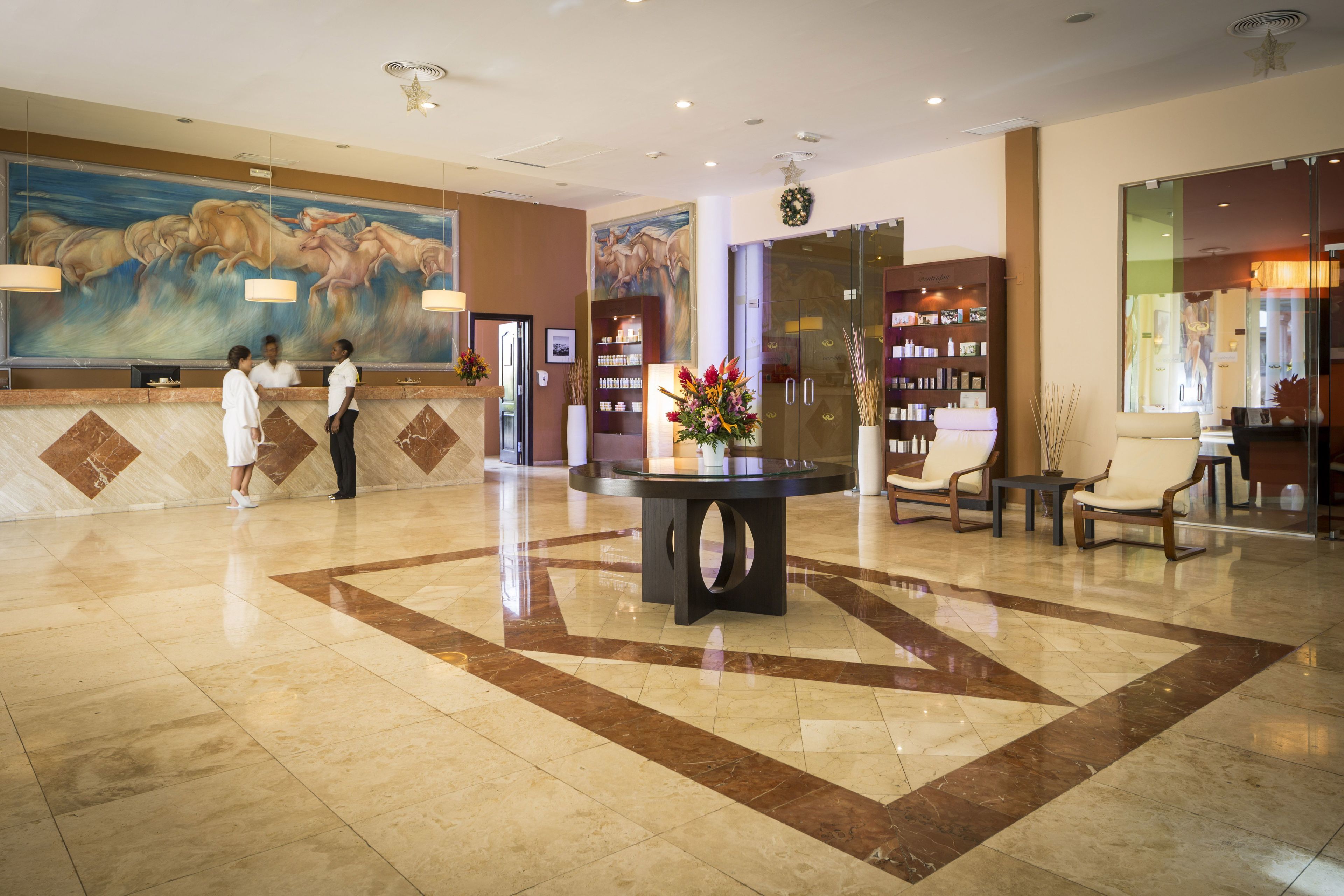 A hotel lobby with a reception desk, a large mural, a round table with chairs, and a glass partition with a view.
