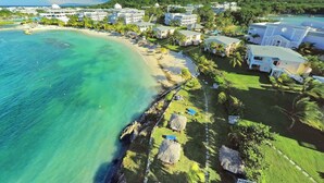 An aerial view of a coastal resort with beachfront villas, clear turquoise waters, and lush greenery.