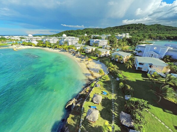 An aerial view of a coastal resort with beachfront villas, clear turquoise waters, and lush greenery.